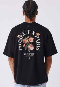 Camiseta de algodón negra con corte oversize. Presenta un gráfico floral colorido y el texto "PROJECT X PARIS WILD SPIRIT PXP COLLECTION" en la parte posterior.