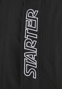 Chaqueta de tela negra con un destacado logo bordado en blanco "STARTER" a lo largo del costado, con textura suave y costuras limpias.