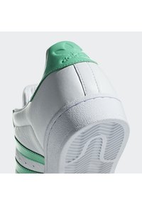 Sneaker blanche avec des accents verts, présentant une tige en cuir texturé, trois rayures vertes et une semelle en caoutchouc avec un motif de semelle.