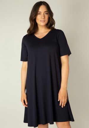 Vrouw met halflang donker haar, gekleed in een knielange, korte mouw, marineblauwe jurk met V-hals, staand tegen een effen achtergrond.