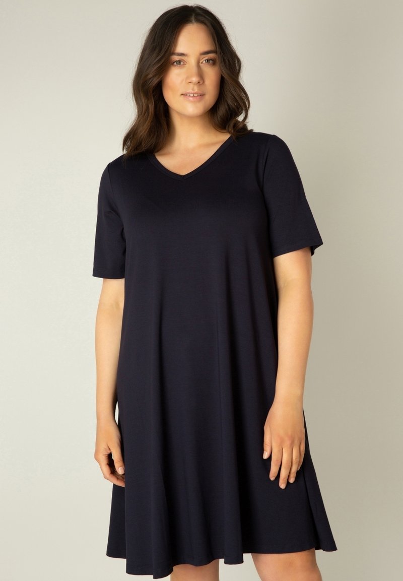 Vrouw met halflang donker haar, gekleed in een knielange, korte mouw, marineblauwe jurk met V-hals, staand tegen een effen achtergrond.