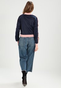 Kvinna som går bort klädd i en marinblå och rosa jacka, högmidjade blå jeans med cropped design och svarta klackboots.