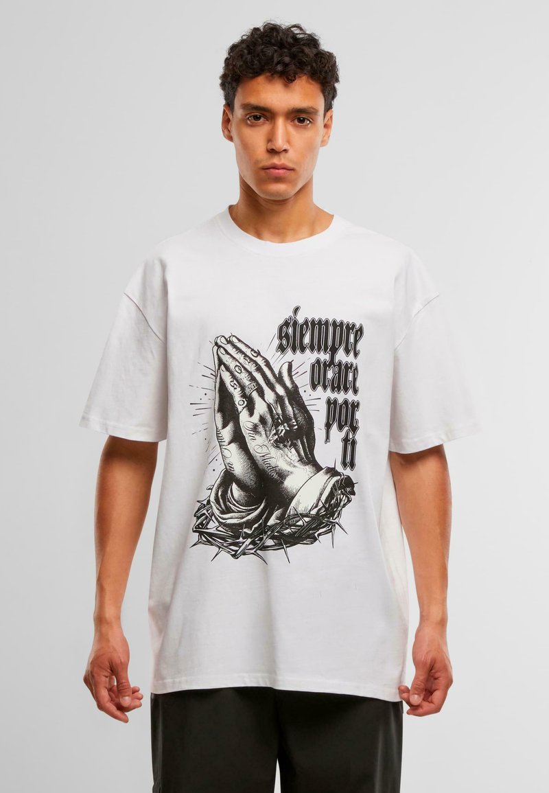 Νεαρός άνδρας φορώντας ένα λευκό T-shirt με μαύρη απεικόνιση προσευχόμενων χεριών και ισπανικό κείμενο "siempre orar por ti" σε ένα απλό φόντο.
