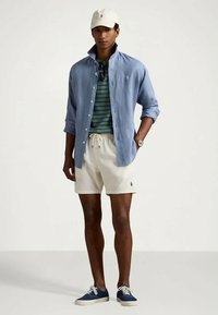 Camisa azul clara de botões sobre uma t-shirt riscada em verde, calções brancos, boné branco e ténis azuis. Apresenta o logo da polo nos calções.