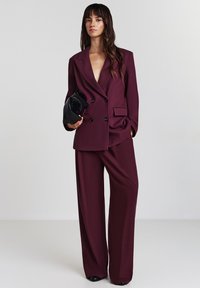 Frau mit langen dunklen Haaren, die einen lockeren burgunderfarbenen doppeltknöpfigen Blazer und weite Hosen trägt, während sie eine schwarze Clutch-Tasche hält.