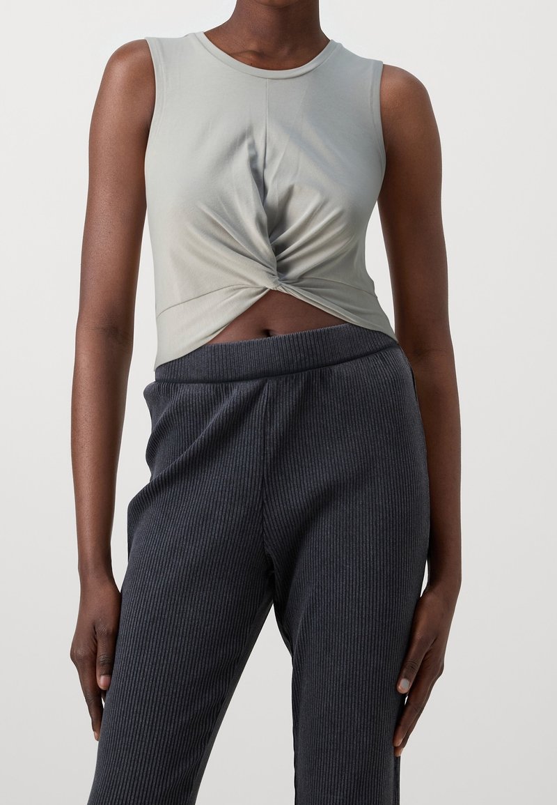 Haut sans manches gris clair avec un détail de devant torsadé, associé à un pantalon côtelé sombre avec une taille élastique.