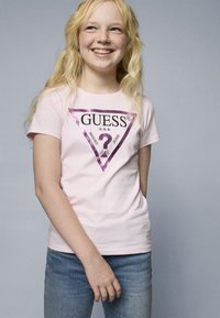 Pink cotton T-shirt featuring a purple triangular logo design with "GUESS U.S.A." text, and "WASHED JEANS" underneath. Worn with blue jeans.