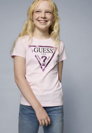 Růžové bavlněné tričko s fialovým trojúhelníkovým logem a textem "GUESS U.S.A." pod ním "WASHED JEANS". Nosené s modrými džínami.