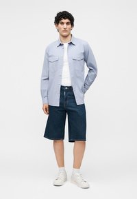 Jeune homme debout, portant une chemise bleu clair boutonnée sur un t-shirt blanc, un short en jean foncé jusqu'aux genoux, des chaussettes blanches et des baskets blanches.
