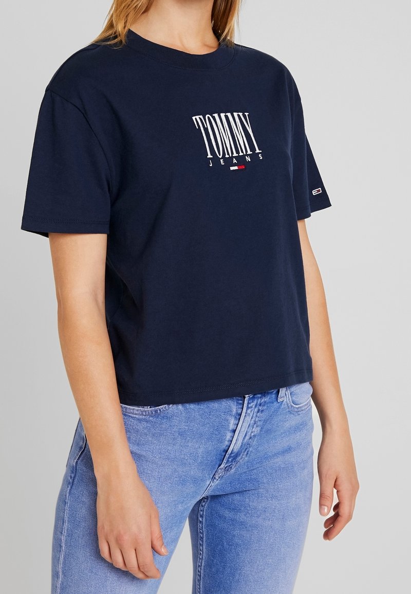 Mörkblå bomulls-t-shirt med korta ärmar, rund halsringning, med vit text och logga "TOMMY JEANS" på framsidan. Avkortad design.