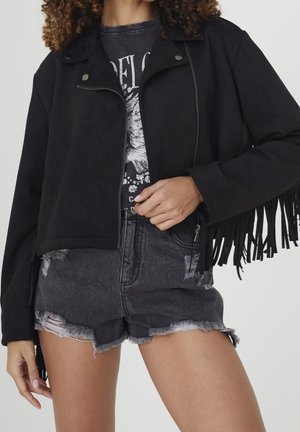 Vrouw draagt een zwarte franjesjas, een grafisch T-shirt en versleten zwarte denimshorts, getoond van schouders tot halverwege de dij.