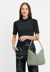 Czarna bluzka krótki rękaw z wysokim dekoltem, zestawiona z błyszczącymi czarnymi skórzanymi spodniami. Modelka trzyma miękką, jasnoszaro-zieloną skórzaną torebkę z paskiem.