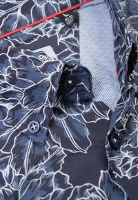 Marineblauw bloes met bloemenmotieven in blauw en wit, knoopsluiting bij de kraag en rode accenten rond de hals. Geweven stof met een gladde afwerking.