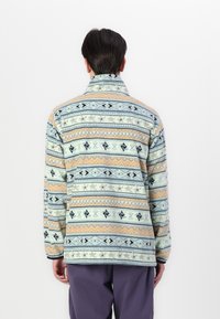 Veste en polaire présentant une base bleu clair avec des motifs géométriques en orange, marine et crème, un col rabattu et de longues manches.