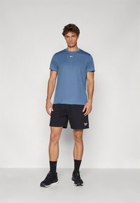 Hellblaues sportliches T-Shirt mit kurzen Ärmeln, kombiniert mit schwarzen Shorts. Schwarze Turnschuhe und Socken vervollständigen das Outfit. Minimalistisches Branding auf dem T-Shirt.