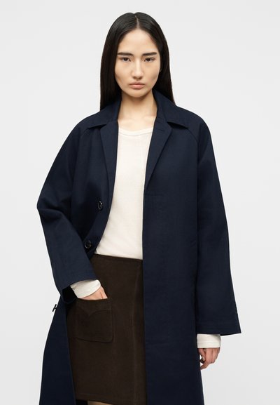 Γυναίκα με μακριά ίσια μαλλιά φοράει μπλε σκούρο παλτό πάνω από λευκό πουκάμισο και σκούρα καφέ φούστα, στέκεται μπροστά σε απλό φωτεινό φόντο.