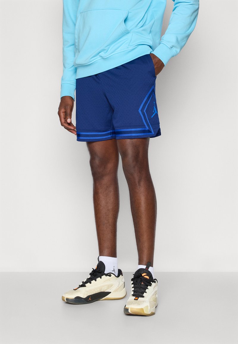 Pantalones cortos deportivos azules con un patrón texturizado, que presentan un logo sutil y rayas en contraste, combinados con zapatillas blancas y negras.