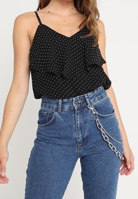 Černý top na ramínka s puntíky a vrstvenými volány, kombinovaný s vysoce pasovými modrými džíny, na kterých je dekorativně připevněný stříbrný řetěz.