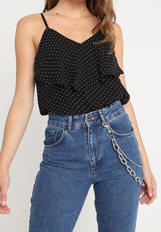 Černý top na ramínka s puntíky a vrstvenými volány, kombinovaný s vysoce pasovými modrými džíny, na kterých je dekorativně připevněný stříbrný řetěz.