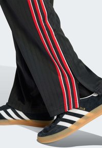 Schwarze Trainingshose mit roten und weißen Seitenstreifen, mit Reißverschlüssen am Saum. Kombiniert mit schwarzen Wildledersneakern mit weißen Streifen und Kautschuksohlen.