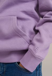 Lavendel sweatshirt med ribbad mudd, kenguruficka och en liten präglad logotyp på ärmen. Mjuk textur och avslappnad passform.
