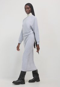 Eine Frau, die ein hellgraues langärmliges Kleid, schwarze Stiefel, einen weißen Rollkragenpullover und einen schwarzen Hijab trägt, steht vor einem schlichten weißen Hintergrund.