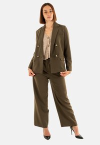Traje entallado en verde oliva con un blazer de doble botonadura, pantalones de pierna ancha y un cárdigan texturizado beige. Tacones altos negros completan el look.