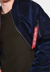 Veste bomber bleu marine à finition brillante, dotée d'une poche zippée et d'une étiquette rouge portant l'inscription « REMOVE BEFORE FLIGHT » attachée au zip.