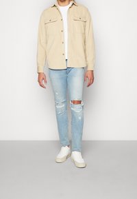 Beige kordflöjtskjorta med två framfickor, bärs över en vit t-shirt, matchad med ljusblå slitna skinny jeans och vita sneakers.