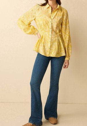 Femme portant un chemisier jaune à manches longues à motifs, associé à un jean évasé bleu et des chaussures tressées marron clair.