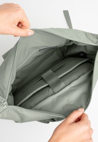 Vihreä kangaskassi, jossa on vetoketjullinen aukko, paljastaen pehmustetun sisätilan, jossa on hihna esineiden kiinnittämiseksi. Sujuva pinta ja minimalistischinen muotoilu.