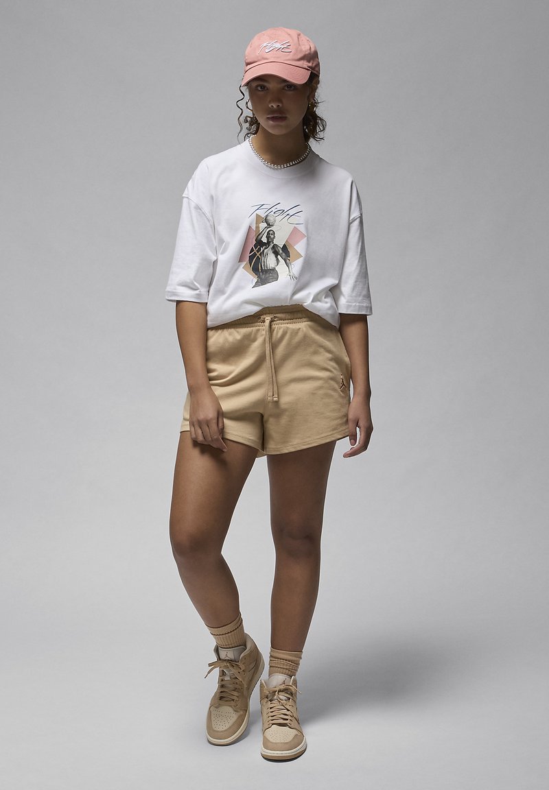 T-shirt bianca con stampa grafica, pantaloni corti beige, cappellino rosa chiaro e sneaker beige. Materiali morbidi, vestibilità rilassata e dettagli delicati.