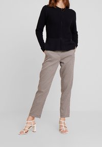 Schwarzer strukturierter Cardigan mit Reißverschluss und glattem Saum, kombiniert mit hellbraunen karierten Hosen und weißen Sandalen mit Blockabsatz und Riemchen.