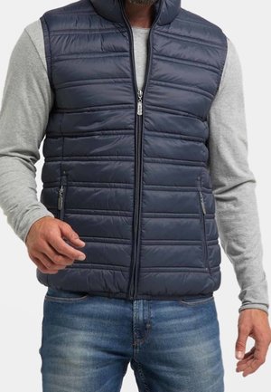 Mężczyzna w granatowej pikowanej kamizelce na zamek, założonej na jasnoszarą koszulę z długim rękawem i niebieskie dżinsy, z lekko uniesionymi rękami.