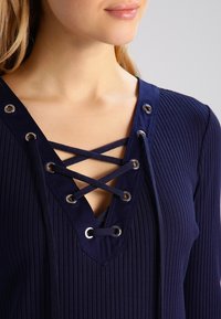 Navy ribbed top with a V-neckline, featuring crisscross lacing and metal grommets. Soft fabric with a textured appearance.