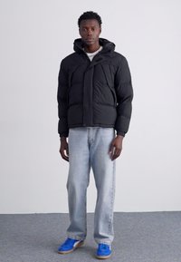 Veste matelassée noire avec capuche, dotée d'une fermeture éclair à l'avant et de plusieurs coutures, associée à un jean bleu clair et des baskets bleues.