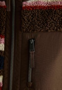 Nahaufnahme einer braunen Jacke aus strukturiertem Fleece-Stoff mit einer senkrechten Reißverschlusstasche und einem Stoff-Ziehriegel.