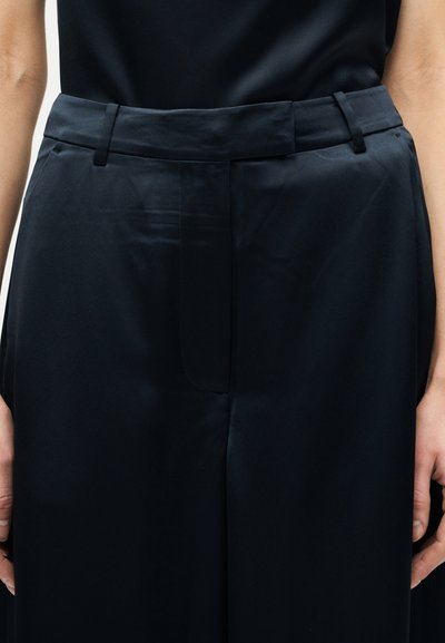 Άτομο που φοράει ψηλόμεσο, σκούρο ναυτικό παντελόνι με θηλιές για ζώνη και πλαϊνές τσέπες, τα χέρια ακουμπισμένα δίπλα στους γοφούς.