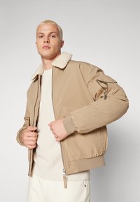 Chaqueta bomber beige con cuello esponjoso, cierre en la parte delantera y bolsillo lateral. Presenta puños y cinturilla acanalados. Llevada sobre un suéter texturizado color crema.