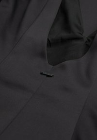 Schwarzes Kleid mit V-Ausschnitt, glatter Stofftextur und einem kleinen Metalletikett, das auf der Vorderseite angenäht ist. Schlichtes, elegantes Design.