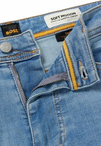 Světle modré džíny z denimu se zipem na předním dílu, žlutým vnitřním páskem a značkou BOSS na štítcích. Vyznačují se kontrastním prošíváním a malým potrhaným místem.