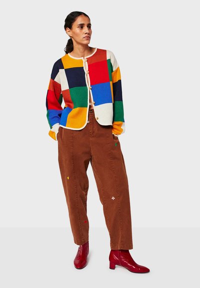 Vrouw met donker haar in een staart, gekleed in een multicolore patchwork cardigan, bruine broek met kleine geborduurde patches en rode enkellaarsjes.