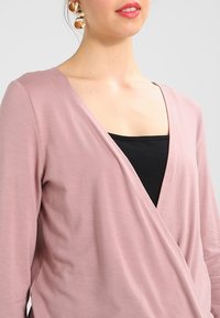 Vaaleanpunainen kääntöpaita, joka on valmistettu pehmeästä kankaasta ja jossa on V-pääntie. Paita on mustan hihattoman paidan päällä, ja siinä on pitkät hihat sekä minimaalista ompelutyyliä.