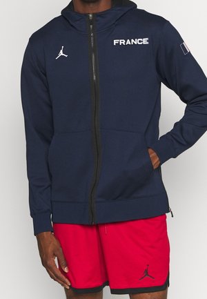Man draagt een marineblauwe hoodie met de tekst "FRANCE" en logo, gecombineerd met rode sportshorts met een klein logo op het linkerbeen.