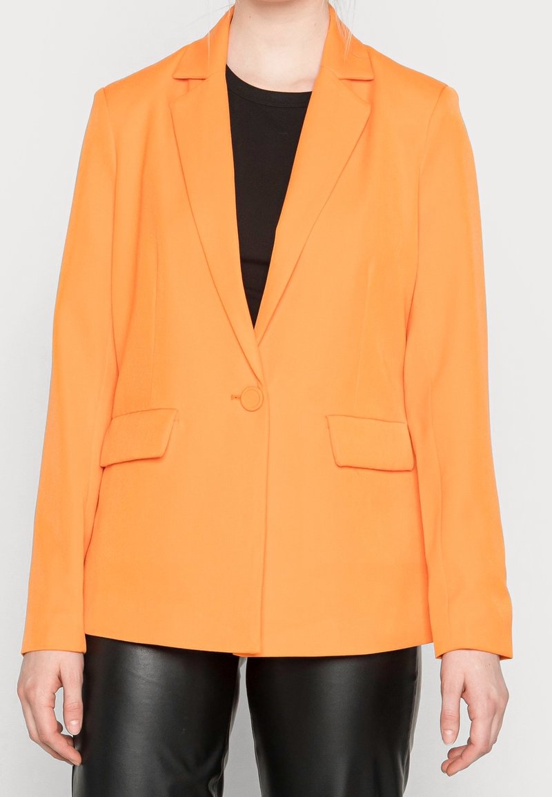 Blazer orange avec revers ouverts, deux poches avant et une fermeture à un bouton. Tissu lisse avec une coupe structurée. Haut noir en dessous.
