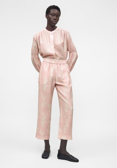 Γυναίκα φοράει ροζ μπλούζα με μακριά μανίκια και μοτίβο παϊέτ, μαζί με ταιριαστό παντελόνι και μαύρα μπαλαρίνια, στέκεται μπροστά σε λευκό μονόχρωμο φόντο.