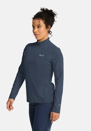 Donna con capelli ricci raccolti che indossa una maglia blu navy a maniche lunghe con cerniera e pantaloni abbinati, in piedi davanti a uno sfondo semplice.