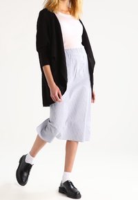 Žena v bílém topu, černém kardiganu, pruhované světle modré sukni, bílých ponožkách a černých botách, stojící na jedné noze na bílém pozadí.