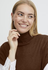 Brun turtle neck sweater med ribbet halslinning, båret over en hvid skjorte med knapper. Stoffet er glat og tætsiddende omkring halsen.