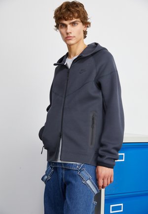 Dunkelgrauer Zip-Hoodie mit zwei seitlichen Taschen und schwarzen Akzenten. Verfügt über eine Kapuze und wird mit blauen Jeans gezeigt. Glatte Textur.